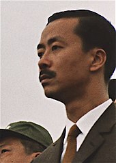Middle-aged man with side-parted black hair and moustache, in a black suit, white shirt and brown tie. To the left is a clean-shaven Asian man with black hair and a green military cap.