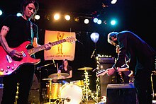 Three people clearly pictured on stage, with a fourth obscured in the background. On the wall in the background there is a blurred poster of the band Carissa's Wierd