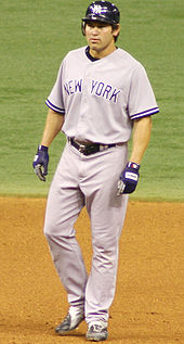 A man in a gray baseball uniform with "NEW YORK" on the chest and a dark batting helmet stands in the infield dirt of a baseball diamond.