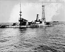 A stripped version of Indiana without gun barrels. The superstructure is seriously damaged and her stacks lean sideways, the front one pointing almost horizontal. A second wreck is visible in the background