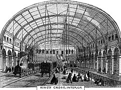 A railway station with a train pulled by an early steam engine. Brick walls rise on both sides and a glass roof arches overhead