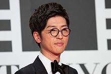 A short black haired Japanese man wearing glasses and black clothes.