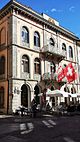 Vue en légère contre-plongée d’une façade d’un bâtiment dans les teintes orangées, munie d’une dizaine de fenêtres en arche.