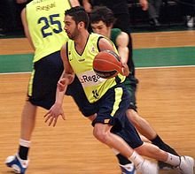 Navarro, en jaune, en mouvement et en dribble. Un défenseur, légèrement en arrière, essaye de le retenir. Au fond de l'écran, un autre joueur en jaune, de dos.