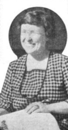 A smiling white woman with short dark hair and eyes tightly closed, wearing a gingham-check dress, hand resting on a braille book