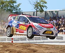 Photographie d'une voiture de rallycross bleue, blanche et rouge, en plein vol, après un saut, de trois-quarts.