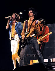 The Rolling Stones on stage in December 1981. From left: Mick Jagger wearing a blue jacket with yellow clothing and a black belt singing into a microphone, Keith Richards wearing black pants and a small purple vest (no shirt) playing a black guitar to the left—and slightly in front—of Jagger, Ronnie Wood wearing an orange jacket and black shirt/pants playing a beige guitar behind Jagger and Richards.