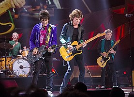 Rolling Stones performing on stage in Milwaukee, Wisconsin. From left: Charlie Watts on brown drum set, Ronnie Wood wearing a purple jacket with black jeans playing a silver coloured guitar, Mick Jagger wearing black shirt and pants playing an orange/yellow guitar, Keith Richards with a green vest and black clothing playing an orange/yellow guitar (similar to Jagger's)