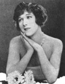 A smiling young white woman with dark hair cut in a bob, hands resting next to her face