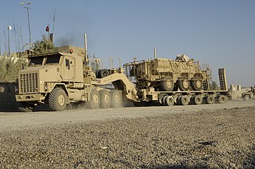 An Oshkosh M1070 8×8 Heavy Equipment Transporter (HET) tractor pulling a 5-bogie M1000 HETS trailer, carrying a slat-armored M93 Fox 6×6 NBC detection vehicle near Baghdad