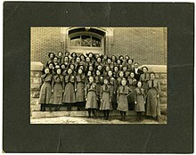 A group portrait of Girl Scouts in Crothersville, Indiana, circa 1945. The girls are gathered in rows and are standing in front of the doors of a brick building.
