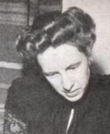 A white woman with hair in a bouffant updo, looking downward, wearing a dark blouse or dress