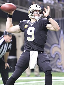 Drew Brees wearing a New Orleans Saints uniform and helmet, winding up to throw a pass with a football in hand.