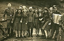 Black and white photograph of about three men in SS uniform and about 10 women in uniform smiling wide on a wooden bridge. One of the men plays an accordion.