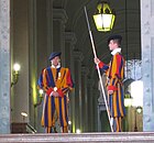 La porte de Bronze, entrée principale du palais apostolique du Vatican, gardée en permanence par deux hallebardiers de la Garde suisse pontificale. À l'intérieur, à gauche, le sergent se tient derrière un bureau. L'Escalier royal est visible au fond, à l'arrière-plan.