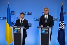 Two men in business suits stand at silver podiums, with a blue and yellow Ukrainian flag to their left and a blue and white NATO flags to their right.