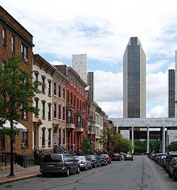 An urban street with cars parked along either side. On the left is a group of ornate three-story brick rowhouses in various colors. In the background is a tall modernist office tower, with smaller versions on its flanks.