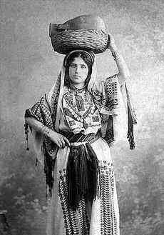 A young woman facing the camera, her left hand holding the edge of a basket with a clay pot inside that is resting on her head, her right hand at her waist. She is looking directly at the camera, wearing a long white dress of heavy material. It is embroidered with geometric designs across the chest (where there is also a v-shaped line of coins) and down the front across the length of her legs. The front part of her hair is parted in the middle, the rest covered with a long veil of the same colour and material as the dress, which is also heavily embroidered. Her arms from the elbows down are not covered by the dress or veil, but are adorned with several bracelets. Around her waist is a belt with fine fringe gathered and hanging down at the center.