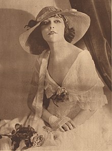 A young white woman with dark hair, seated, wearing a hat with a wide trim and a white gown