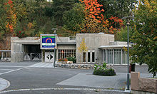 Photographie de l'entrée d'un bâtiment en béton, avec une place goudronnée en premier plan et des arbres en arrière-plan.