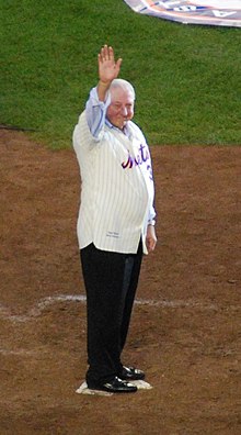 A white-haired man wearing a white pinstriped baseball jersey with "Mets" across the chest and dark slacks stands on home plate.