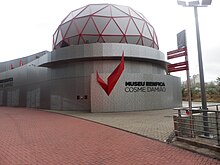An outside view of the Benfica Museum. Its façade consists of grey metal panels with a red wing- or eagle-shaped piece of art followed by "MUSEU BENFICA COSME DAMIÃO" in black letters. On the top of the building, there is a red geodesic dome with a grey covering.