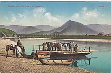 Carte postale montrant une barque à Tchouïozy sur la Katoun avant la construction du pont d'Inya. En arrière-plan se trouvent un paysage montagneux verdoyant, ainsi que quelques maisons en bois sur l'autre rive. Cinq personnes et trois chevaux sont déjà sur la barque, tandis que quatre autres sont sur la berge avec leurs deux chevaux. Il y a aussi deux petites pirogues à côté de la barque.