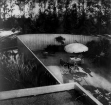 An outdoor patio in Ayn Rand and Frank O'Connor's San Fernando Valley home, photographed from above. In the center of the image is a table with an umbrella over it; notice that a man who may be Frank O'Connor sits on the left side of it. Ayn Rand sits slightly away from the table on the right side. There is lush vegetation surrounding and throughout the patio.