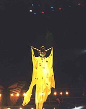 Velasquez performing in a long yellow gown while hanging on a harness