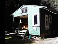 A green hut with a 4 steps up to a small porch. Lots of small windows including one above the porch like a house truck might have. Trees surround the hut..