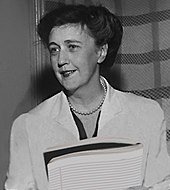 Black and white portrait of a woman with short hair, standing in front of a tartan backdrop, wearing a white suit with a pearl choker and holding a report in both hands in front of her torso.
