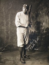 Full body shot of baseball player Babe Ruth, holding a bat and wearing a "NY" hat.