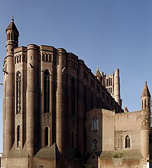 photo couleur d'une cathédrale en brique rouge vue depuis un côté. Le bas taluté est massif. Le haut est marqué par l'opposition de tourelles inclues dans le mur et de hautes et fines fenêtres à vitraux. En arrière-plan, le clocher dépasse de la toiture.