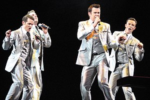 Four adult males are singing into hand-held microphones. They all wear silver-grey three-piece suits, white shirt and dark tie. The man on the left is shown in mostly right profile and partly obscures the second man. Both hold the microphone in their left hands with the right hand raised in a clenched fist near their shoulder. The next two men hold the mics in their right hands and raise their left as a fist.