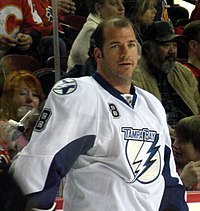 Hockey player in white Tampa Bay uniform, with his helmet off. He looks toward the camera, and has dark hair with a receding hairline.