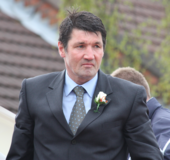 Head and upper torso of a middle-aged dark-haired white man dressed in a smart suit with floral buttonhole.