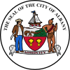 Circular seal with central images of a shield at center and sailing ship above it, with a European man to the left and a Native American to the right. The seal's edge reads "THE SEAL OF THE CITY OF ALBANY" with "ASSIDUITY" in a banner above the bottom.