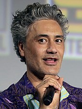 A man with black and silver slicked-back hair wears a purple patterned shirt and holds a microphone.