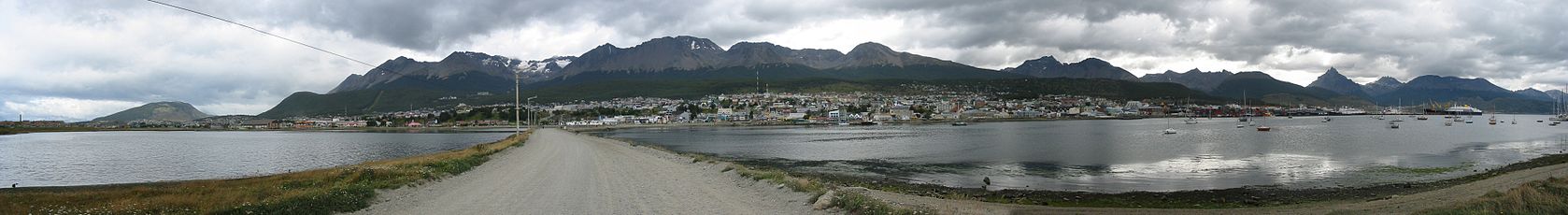 A small city across a gray waterway under lowering gray clouds. A road leads to the city across a causeway. Mountains with snow and a low treeline form the backdrop. A few boats are in the water.