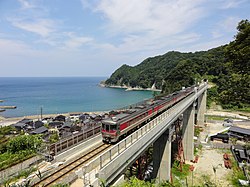 Diesel multiple unit train crossing the concrete Amarube Viaduct. A green hillside is visible on the right, and a bay of the Sea of Japan on the left.