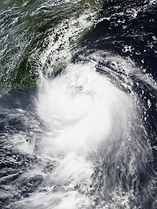 A photograph of a tropical storm over the western Pacific Ocean. The storm possesses an eye-like feature, but convection is limited on the northern half of the circulation; instead, a large area of thick, deep convection is sprawling over the storm's southern half. The overall cloud pattern is tight and well-organized.