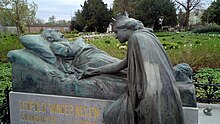 Fotografie. Im Vordergrund ist der Grabstein von Leopold Vander Kelen zu sehen. Der Grabstein ist eine Statue von zwei Personen. Sie zeigt den aufgebahrten Vander Kelen auf seinem Totenbett, die Arme vor der Brust verschränkt. Vor Vander Kelen kniet eine gekrönte Person, die einen Zweig neben den Toten legt. Der Sockel trägt die Inschrift „LEOPOLD VANDER KELEN“ und darunter „BOURGMESTRE DE LOUVAIN“. Im Hintergrund sieht man den Friedhof von Löwen.