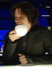 Guy Sigsworth looking towards his right and smiling, while drinking from a cup in his right hand.