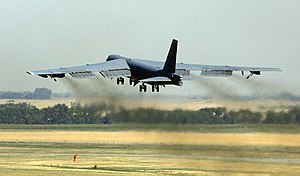 A large, grey, jet transport plane is taking off away from the camera, with the wash from the jets blurring the landscape between the aircraft and the ground.