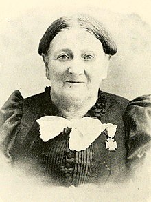 Older white woman wearing a medal on her black dress.