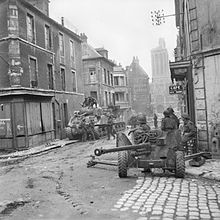 An intersection; an anti-tank gun is covering the crossroads in the foreground and in the street behind are some soldiers and two tanks