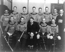 A black and white photo of ice hockey players sitting in two rows on a bench. Their jerseys feature the American flag on the front. Their coach is seated front row, center, with a young boy sitting in front of him.