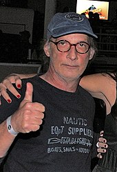 A man with glasses is wearing a black shirt and cap with a thumbs-up pose