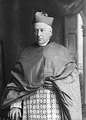 A man wearing a mozzetta, liturgical vestments, and pectoral cross faces forward.