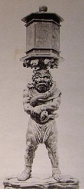 Front view of a stocky statue with a demon face. He is balancing a lantern on his head. He is grabbing his right wrist with the left hand. Black and white photograph.